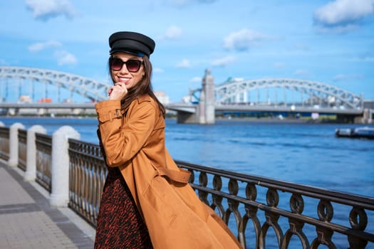 Beautiful young woman of caucasian appearance in a brown raincoat and dress posing on the embankment on a sunny day