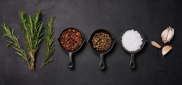 Miniature pans with spices, salt, black pepper and fragrant pepper, a sprig of rosemary on a black table. Spices for cooking