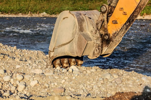 Yellow digger (excavator machine) bucket digging in stones ground by the river.