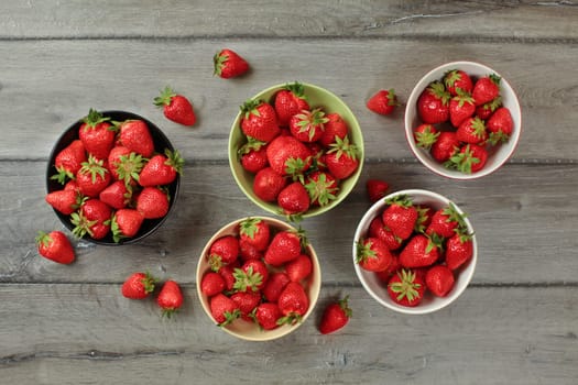 Tabletop view, small bowls with strawberries, some of them spilled on gray wood desk.