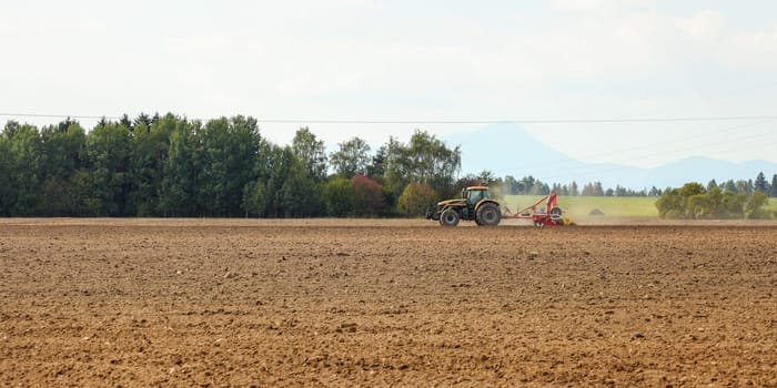 Tractor sowing in empty field on countryside with some mountains in background.