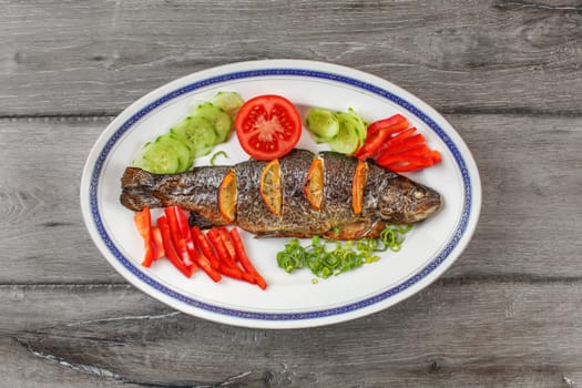 Tabletop view, grilled trout on oval plate, orange slices, tomatoes, pepper, cucumber and green onion around.