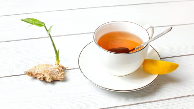Porcelain cup with hot amber tea, and slice of lemon, dry ginger root green sprout text on white boards desk.