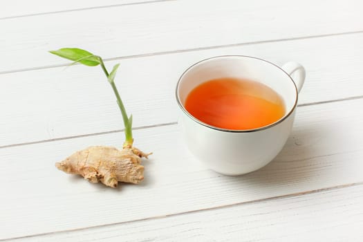 White porcelain cup with hot amber tea, dry ginger root and green sprout next to it on white boards desk.