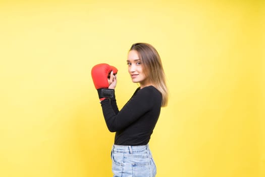 Portrait of young woman in a casual clothes and hands in boxing gloves against yellow background