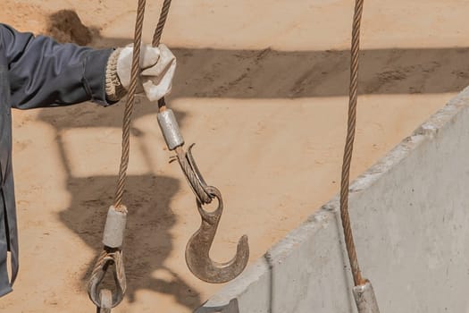 The hand of construction worker male take a heavy iron hook lifting mechanism with a steel cable rope to a concrete, cement structure.