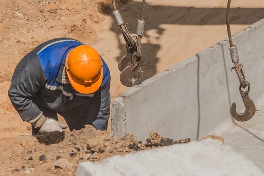 An industrial worker in overalls and a protective helmet attaches a heavy iron hook lifting mechanism with a steel cable rope to a concrete, cement structure.