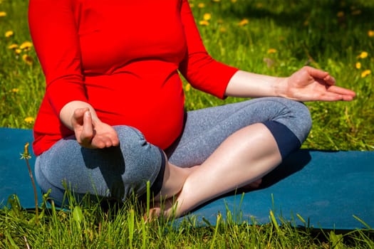 Pregnancy yoga exercise - pregnant woman doing asana Sukhasana easy yoga pose with chin mudra outdoors on grass lawn with dandelions in summer