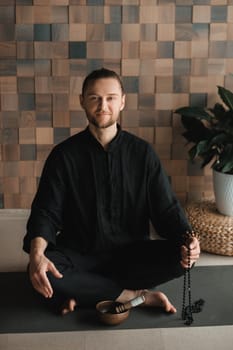 Portrait of a young man in a black kimano sitting in a lotus position on a gym mat in the interior.