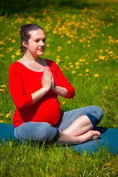 Pregnancy yoga exercise - pregnant woman doing asana Sukhasana easy yoga pose with namaste salutation outdoors on grass lawn with dandelions in summer