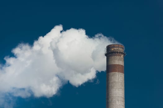 Pollution of the environment, ecology and air. Withdrawal of combustion products of soot, smoke and gases from the pipe of an industrial plant into the atmosphere against the background of a blue sky.