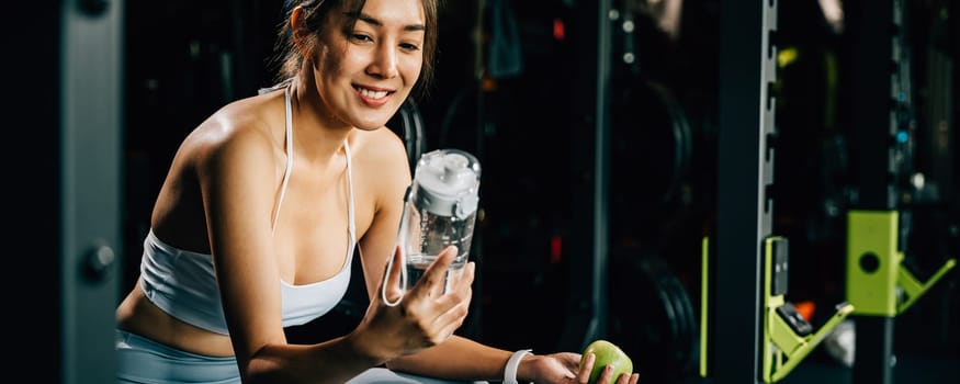 Asian woman holding a green apple and a water bottle in a fitness gym, reminding viewers of the importance of a balanced diet and hydration for achieving a fit and healthy body. healthy lifestyle