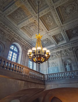 Architectural details of a ornate staircase hall with a glowing vintage chandelier hanging from ceiling at Versailles Palace, France