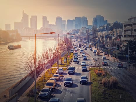 Busy traffic on highway and city streets creating high pollution and smog. Urban sunset view across Seine river to La Defense metropolitan district, Paris, France