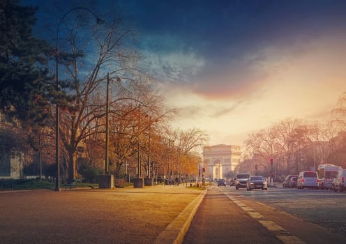 Triumphal Arch (Arc de triomphe) in Paris, France. The famous historic landmark in sunset light seen from the city street with busy traffic