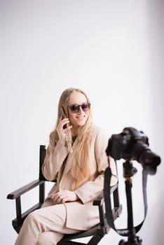 A woman producer videographer, a blonde assistant with a camera and smartphone, happy talking on the phone and smiling in the studio. Wearing a formal nude pantsuit on a white isolated background. Verical