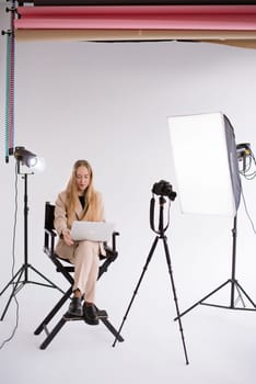 A business woman videographer , assistant gafer in a suit is working, typing on a laptop MacBook and sitting on a producer chair. Video Production studio, tripod camera , soft box , light