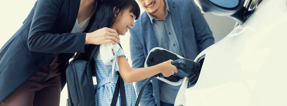 Progressive young parents and daughter with electric vehicle and home charging station. Green and clean energy from electric vehicles for healthy environment. Eco power from renewable source at home.