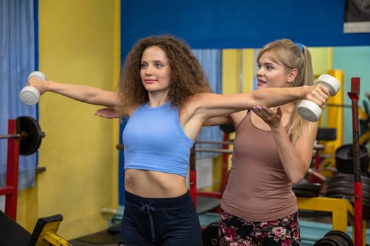 Two female friends are working out at the gym. One woman stands in front and lifts weights in her upright hands. The other woman stands in the back and supports her friend's arms while belaying her.