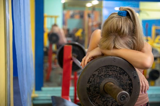 Young woman at the gym. A woman exhausted from an intense workout at the gym. She leans against an exercise barbell. The woman hides her face in her hands leaning against the barbell. Back blurred view of large mirror hanging on wall.