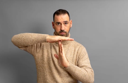 Bearded Hispanic man in his 40s wearing a beige turtleneck making the time-out gesture while looking seriously at the camera, isolated on gray studio background