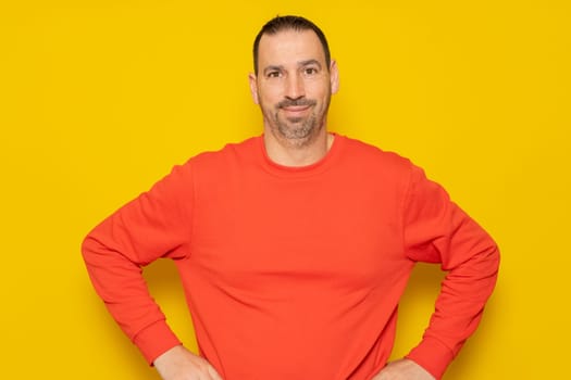 Cheerful bearded man wearing a red sweater posing isolated on gray background, studio portrait. People lifestyle concept. Mock up copy space. Standing with arms akimbo on waist blinking