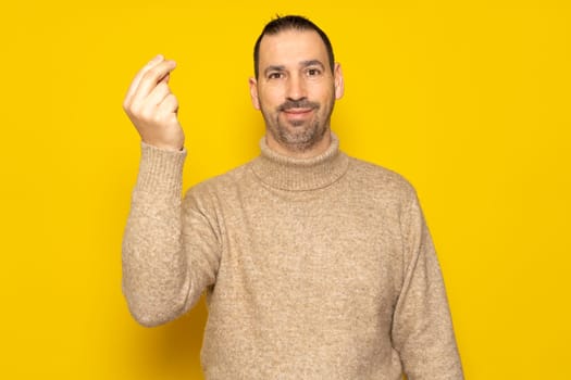 Bearded Hispanic man wearing a beige turtleneck making an Italian mama mia gesture while smiling at the camera, isolated over yellow background.