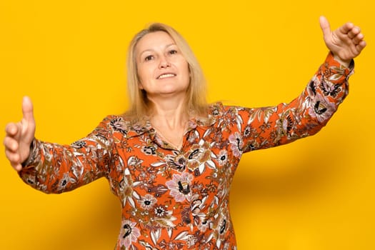 Caucasian blonde woman in a patterned dress with her arms outstretched ready to give a loving hug, isolated on yellow background.