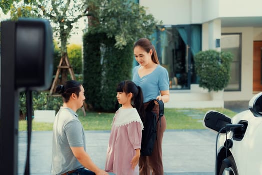 Progressive young parents and daughter with electric vehicle and home charging station. Green and clean energy from electric vehicles for healthy environment. Eco power from renewable source at home.