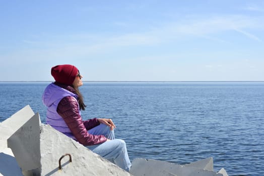 A young woman in a hat on a sunny spring day sits on the breakwaters and admires the sea. Girl in warm clothes on the breakwater