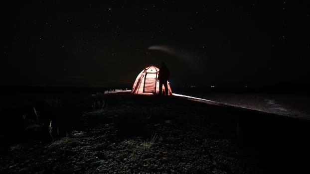 The guy is standing near a tent with a stove at night. Stars and constellations are visible. A lantern is burning in the tent, a pipe from a fresh camp stove is smoking. Bushes grow in places