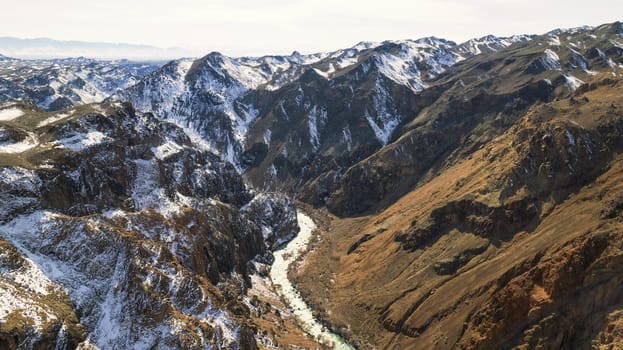 A long river with green water in the Charyn Canyon. There is white snow in places. Bushes and trees grow. The river runs through a canyon among rocks and cliffs. Mountains are visible. Drone view