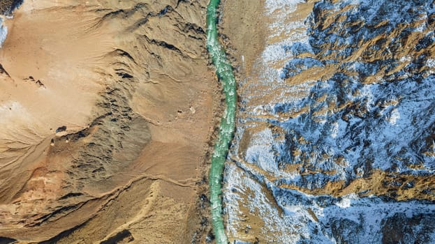 A long river with green water in the Charyn Canyon. There is white snow in places. Bushes and trees grow. The river runs through a canyon among rocks and cliffs. Mountains are visible. Drone view