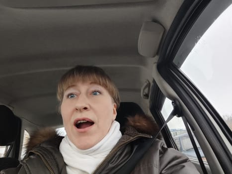 Portrait of adult cute funny blonde woman in warm clothes taking a selfie inside the car and driving it. The concept of pleasant trip in spring, autumn, winter