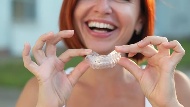 Red-haired Caucasian woman holding transparent mouthguards for bite correction outdoors. A girl with a beautiful snow-white smile uses silicone braces.