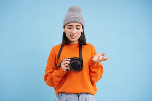 Portrait of asian woman in hat, holding digital camera with confused face, unprofessional photographer doesnt know how to take pictures on digicam, blue background.