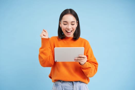 Cheerful young woman with digital tablet, looks happy at gadget and celebrating, laughing and smiling, standing over blue background.