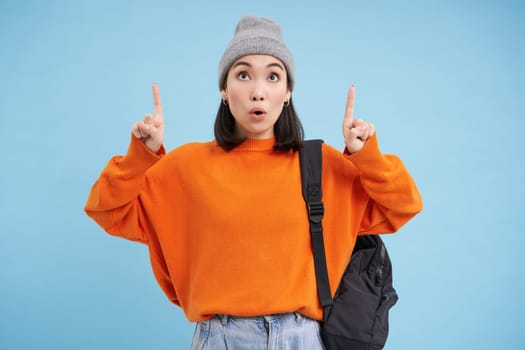 Portrait of asian girl with shocked face, pointing fingers up and say wow, drops jaw, looks impressed, stands over blue background.