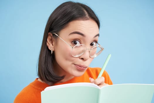 Portrait of cute korean woman in glasses, holding pen and notebook, student making notes, tutor teaching lesson, blue background.