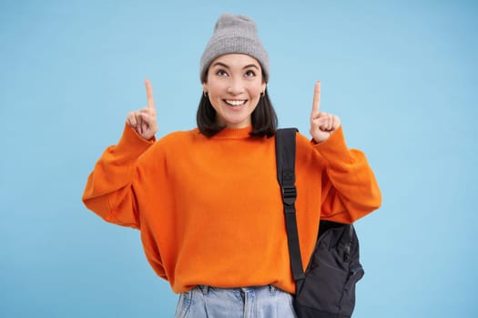 Smiling beautiful korean woman, pointing finger up, shows advertisement on top, wears street outfit and backpack, blue background.