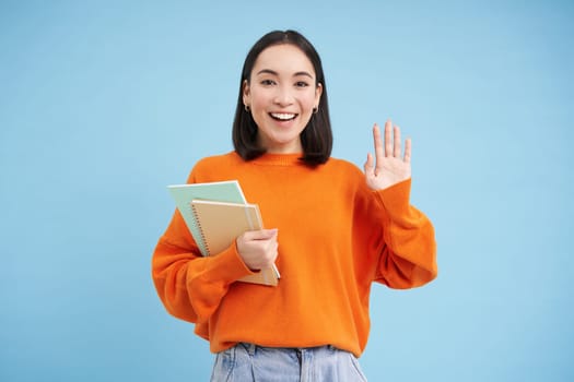 Friendly young woman, student says hello, stands with notebooks and waves hand to greet you, stands over blue background.