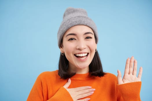 Close up of cute korean girl in beanie, raises one hand and introduces herself, greets you, smiles and looks friendly, blue background.