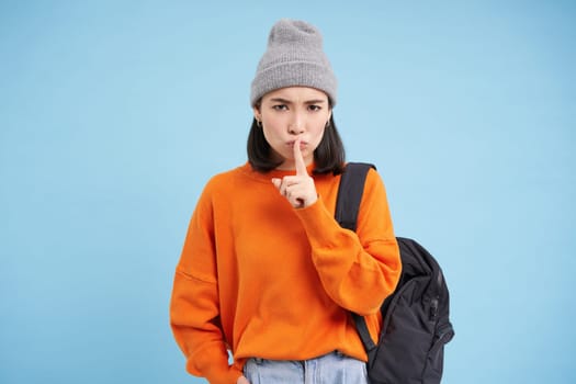 Asian girl shows shush, shh gesture, puts finger over lips, keep quiet sign, stands over blue background with backpack and wears hat.