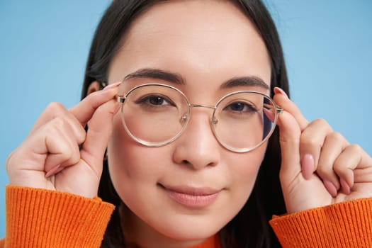 Close up portrait of amazed girl, looks closer at camera in glasses, standing against blue background. Copy space