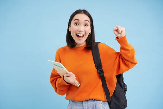 Enthusiastic girl student, celebrating, wearing backpack and holding notebooks, college homework, standing over blue background.