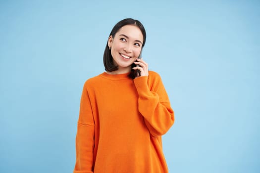 Portrait of young candid woman, asian girl talking on cellphone, holds mobile phone and speaks, stands over blue background.