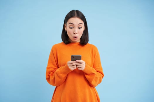 Young woman looks amazed at her cellphone, reads exciting news, found good deal online on mobile application, standing over blue background.