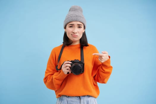 Asian girl in hat, points at her digital camera with disappointed, upset face, doesn't like her device, photographer complains at her digicam, blue background.