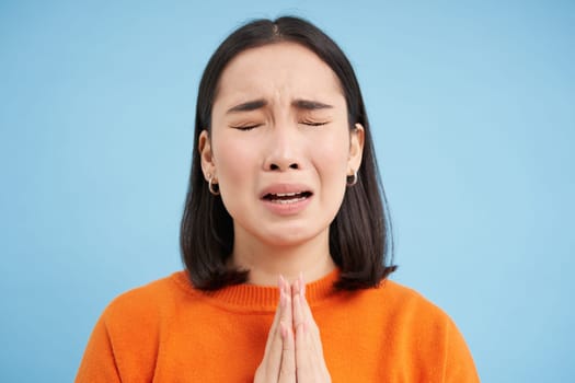 Close up of sad young asian woman, holds hands in pray, begging, pleading for help, stands over blue background.