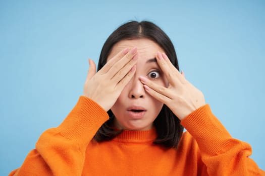 Close up portrait of korean girl waits for surprise, shuts her eyes and peeks through fingers with excited smile, stands over blue background.